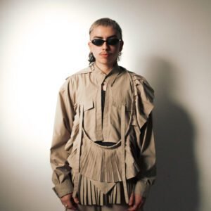 Camisa de tablas “Travesía” en tono beige, hecha de uniformes mineros rescatados. Diseño estructurado con pliegues profundos y detalles en capas. Modelo con lentes oscuros en fondo neutro.
