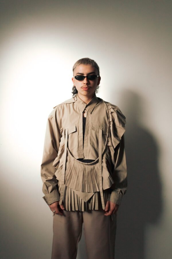 Camisa de tablas “Travesía” en tono beige, hecha de uniformes mineros rescatados. Diseño estructurado con pliegues profundos y detalles en capas. Modelo con lentes oscuros en fondo neutro.