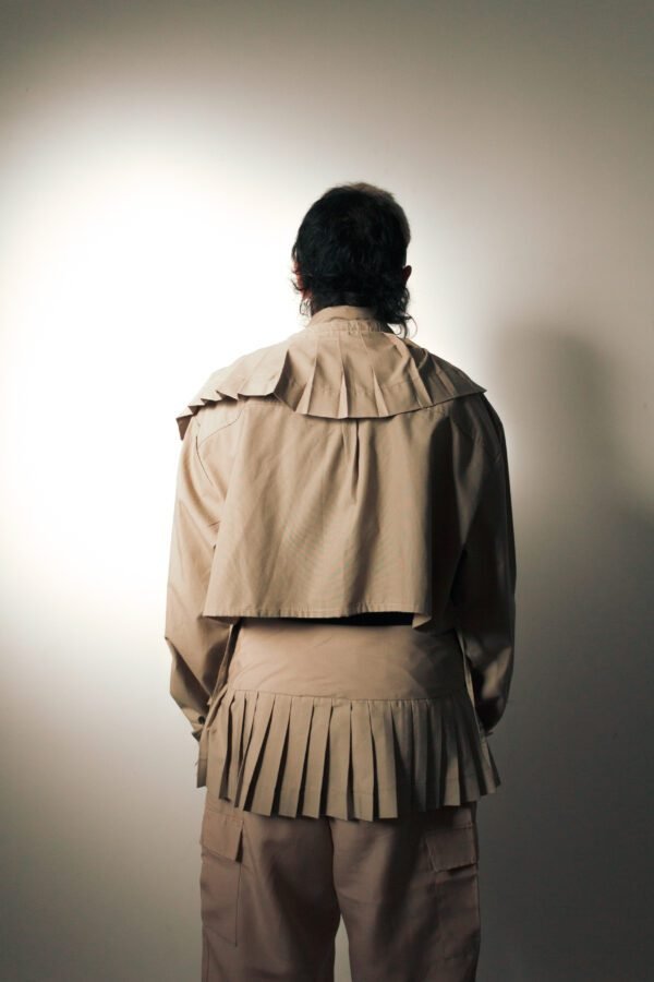 Camisa de tablas “Travesía” en tono beige, hecha de uniformes mineros rescatados. Diseño estructurado con pliegues profundos y detalles en capas. Modelo con lentes oscuros en fondo neutro.